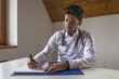 © GENETTICA - Young focused doctor writing a report in his office. He is sitti