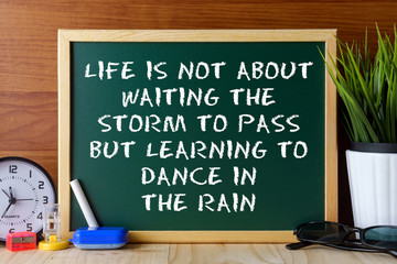 Word quote LIFE IS NOT ABOUT WAITING THE STORM TO PASS BUT LEARNING TO DANCE IN THE RAIN written on green chalk board on wooden table.