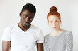 © WHstudio Leushin N - Portrait of young Caucasian female with red hair, angry and disappointed with her African boyfriend, who is standing next to her, looking at the camera with tired expression, fed up with quarrelling