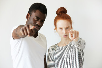 Wall Mural - A couple of youngsters pointing index finger in reproach at camera. Mix of Caucasian and African complexions makes this photo looks rich in contrast. Blame and guilty concept. Negative emotions.