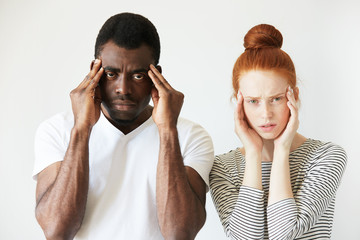 Wall Mural - Interracial relationships concept. Black man in white polo shirt and his Caucasian redhead wife, having headache, holding hands on their temples, looking at the camera, exhausted with endless quarrels