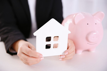 Wall Mural - Savings concept. Woman holding piggy bank in one hand and house figure in another, close up