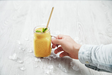 Man hand takes transparent rustic jar with tasty healthly freshly made yellow smoothie from mango, pineapple, passion fruit. Melted ice around isolated on white table