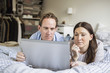 © Maskot - Couple using laptop while lying in bed at home