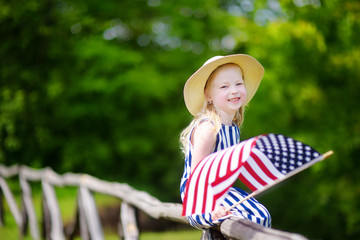 Adorable little girl wearing hat holding american flag outdoors on beautiful summer day