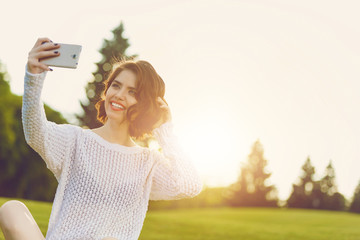 Wall Mural - Outdoor photo of young woman sitting on trimmed green grass field with cell phone. Beautiful tender woman with red hair posing in summer park.