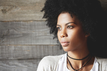 Highly-detailed portrait of pretty African young woman with clean healthy skin and Afro haircut, looking away with thoughtful expression, dreaming of travelling or future plans against wooden wall