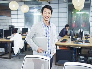 Wall Mural - portrait of a young asian entrepreneur standing in office with laptop under arm