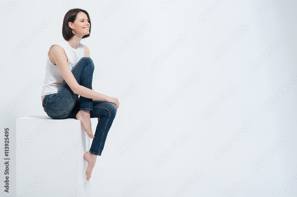 Beautiful young woman sitting on white cube in studio - obrazy, fototapety, plakaty 