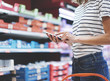 Young woman shopping healthy food in supermarket blur background. Close up view girl buy products using smartphone in store. Hipster at grocery using smartphone. Person comparing the price of produce.