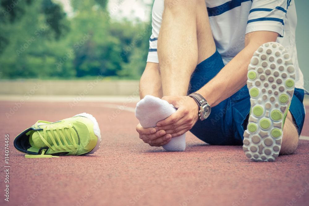 Jogger hands on foot. He is feeling pain as his ankle or foot is broken or twisted. Accident on running track during the morning exercise. Sport accident and foot sprain concepts. - obrazy, fototapety, plakaty 