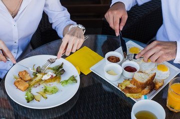 Closeup of business people hands, male and female having breakfast at outdoors cafe. Meals with salad, omelette, bacon.