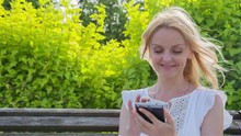 High Quality 10bit Footage Of Happy Smiling Girl Using A Smart Phone In A City Park Sitting On A Bench. Made From 14bit RAW.