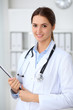© rogerphoto - Young brunette female doctor standing with clipboard and smiling at hospital.  Physician ready to examine patient