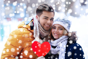 Wall Mural - Young couple in love holding red paper heart outdoors in winter. Snow effect