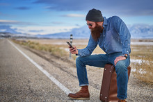 Bearded Hitch Hiker Sitting On Suit Case Using Smart Phone