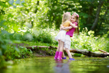 Wall Mural - Two cute little sisters playing in a river wearing raining boots