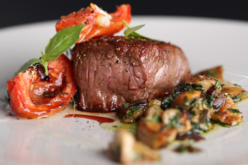 Roasted meat with tomatoes and mushrooms on a white plate, dark background in the restaurant. Close up.