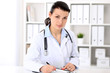 © rogerphoto - Young brunette female doctor sitting  with clipboard near window in  hospital and filling up medical history form.