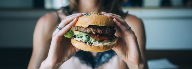 Young girl holding in female hands fast food burger, american unhealthy calories meal on blue background, mockup with space for text message or design, hungry human with grilled hamburger front view