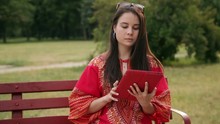 Beautiful Asian Girl In Red Dress Is Sitting On The Bench In The City Park And Looking At The Screen Of A Tablet