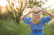 © goodmoments - Back view on blond woman in beautiful blue dress holding straw hat in her hands and having fun in the park outdoors. Girl walking in the park on sunset. Lifestyle and summer concept.