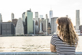 Fototapeta Miasta - Young woman in striped shirt looking at Manhattan skyline at Brooklyn bridge park during sunset