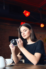 Wall Mural - Portrait of young teenager photographer with long hair in black t-shirt in cafe. Beautiful girl holding old analog film camera in her hands and looking at viewfinder