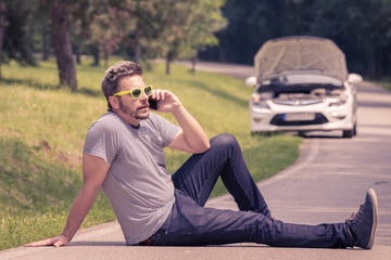 Young modern man sitting on the asphalt and making call with road assistance service. Car with raised hood at the roadside.