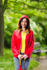 Wall Mural - Portrait of a beautiful young woman in hooded red raincoat looking at camera and smiling while walking in the park on a rainy day.