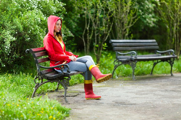 Wall Mural - Shot of a beautiful young woman in hooded red raincoat and rubber boots holding red umbrella. She is sitting on a bench in the park on a rainy day and looking away.