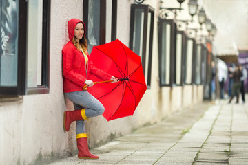 Wall Mural - Portrait of a beautiful young woman in a red hooded raincoat leaning on the wall and looking at camera. She is holding red umbrella.