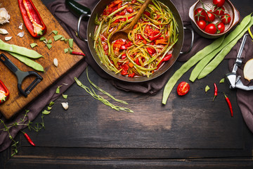 Wall Mural - Green beans with tomatoes sauce in cooking pot with ingredients on cutting board for vegetarian dish on dark rustic wooden background, top view