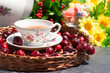 Чай из ягод в изящной чашке на плетеном подносе. Вишни вокруг напитка создают аппетит. На заднем плане стоит ваза с конфетами и лежат цветы.