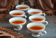 Чай налитый в шесть изящных кружек на фоне коричневых бархатных штор. Напиток подготовлен к чаепитию.
