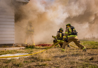 Fire fighters stand by with a hose while another fire fighter battles through heavy smoke to break open a window to allow water to be sprayed on the fire inside. 