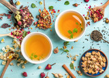 Fototapeta Uliczki - Two cups of healthy herbal tea with mint, cinnamon, dried rose and camomile flowers in spoons over blue background