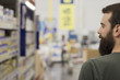 © GENETTICA - Bearded young man in a supermarket looking at the products on a shelf on one side.