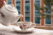 Женская рука держит заварочный чайник и наливает чай в чашку на балконе. Бежевое полотенце и нежный рисунок сервиза создают хорошее утреннее настроение.