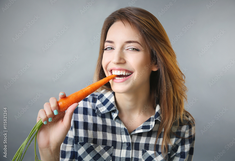 Beautiful girl eating carrot on grey background