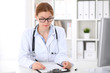 © rogerphoto - Young brunette female doctor sitting at the table and working at hospital office.  Health care, insurance and help concept. Physician ready to examine patient