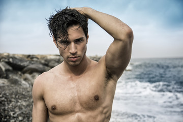 Attractive muscular young shirtless athletic man standing next to water by sea or ocean shore, looking at camera in a cloudy summer day