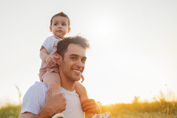 Beautiful portrait of young attractive smiling father with daughter toddler, baby on his shoulders on sunset nature background.