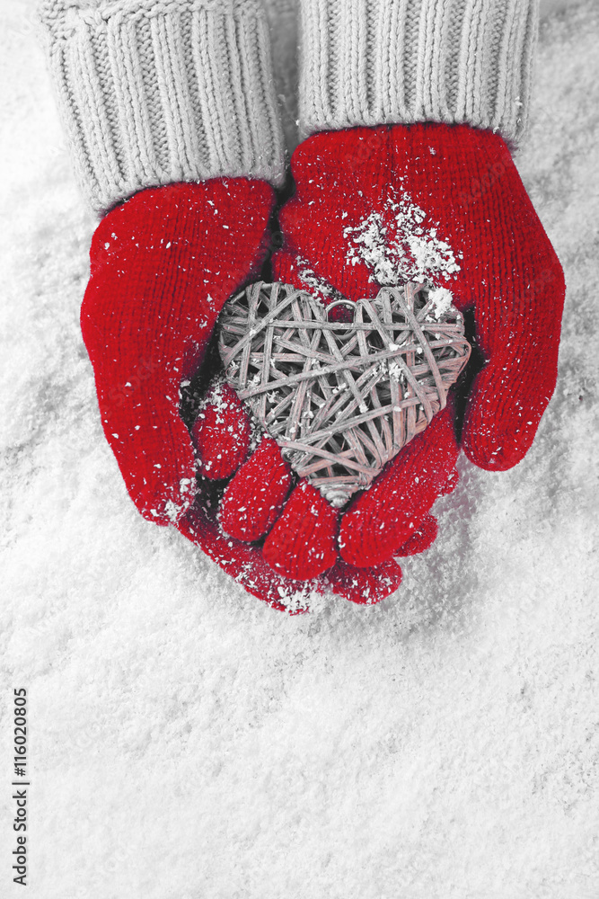 Hands in warm red gloves holding wicker heart on snowy background