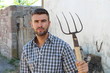 Portrait of young bearded handsome farmer in casual checkered shirt with old pitchfork on rustic background
