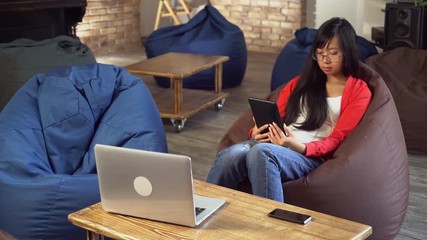 Poster - Young asian woman reading ebook in loft room. Girl spends free time at home in casual interior. On the coffee table laptop. Female holding touch screen tablet with app for read. Student brunette