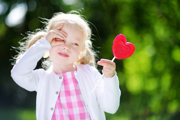 Cute little girl eating huge heart-shaped lollipop outdoors