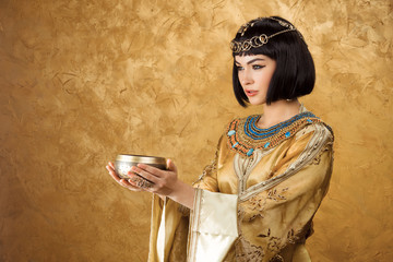 Beautiful woman like Egyptian Queen Cleopatra with cup on golden background