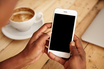 Cropped shot of black female's hands holding mobile phone with blank copy space screen for your text message or promotional content. Young woman watching videos using cell phone sitting at cafeteria