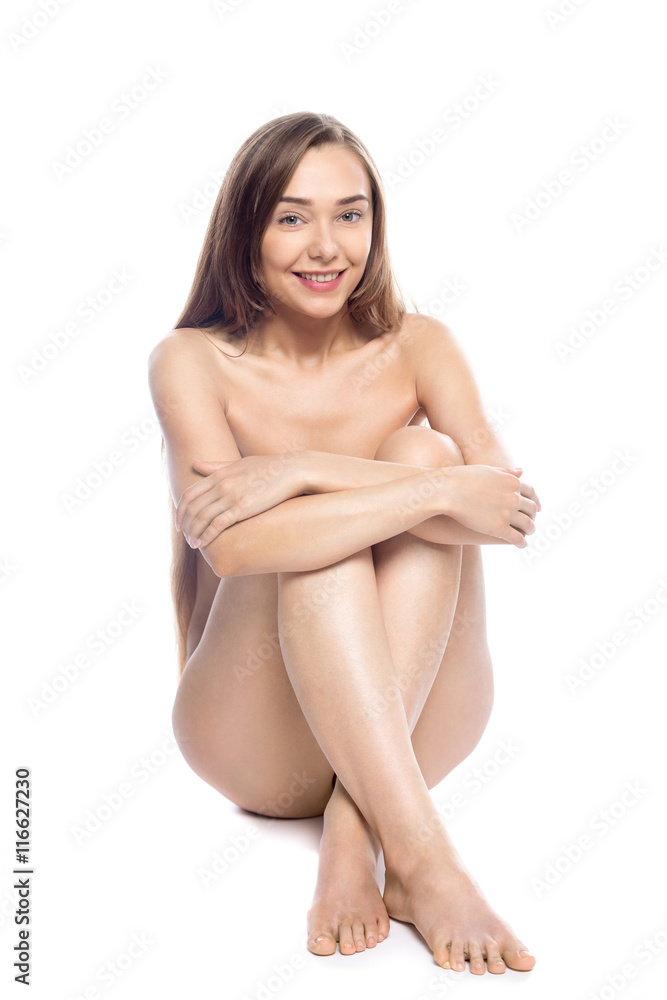 Portrait of young happy slender naked beautiful Caucasian model with long legs sitting against white background. Pretty person posing nude with crossed perfect legs. Studio isolated shot. Full length